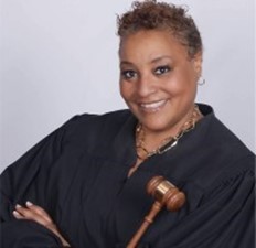 A black women is smiling and wearing a black robe while holding a gavel with her arms crossed